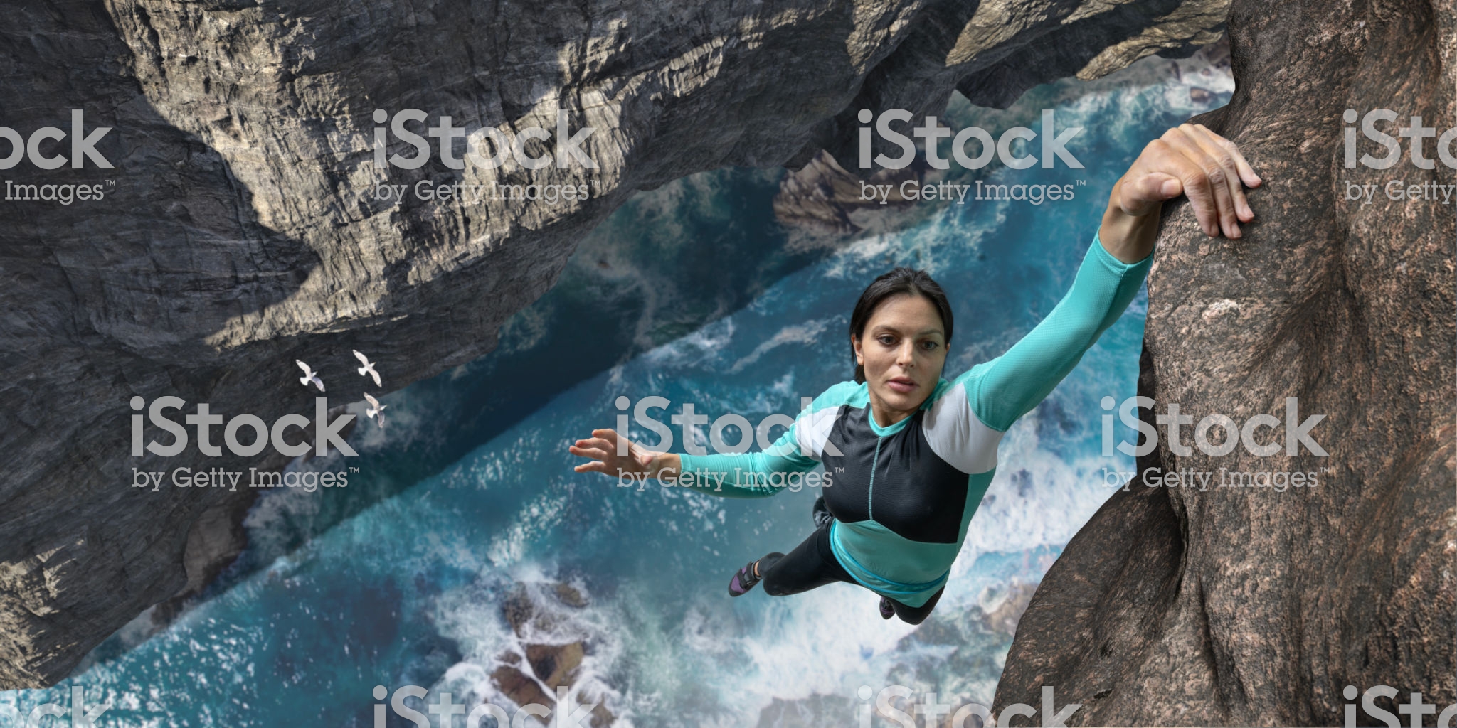A young woman extreme free climbing without safety equipment, hangs one handed from a rock face over the sea and rocks below. The climber is wearing climbing top, leggings. chalk bag and climbing shoes. Waves break over rocks in the sea below during sunny daylight in clear weather.